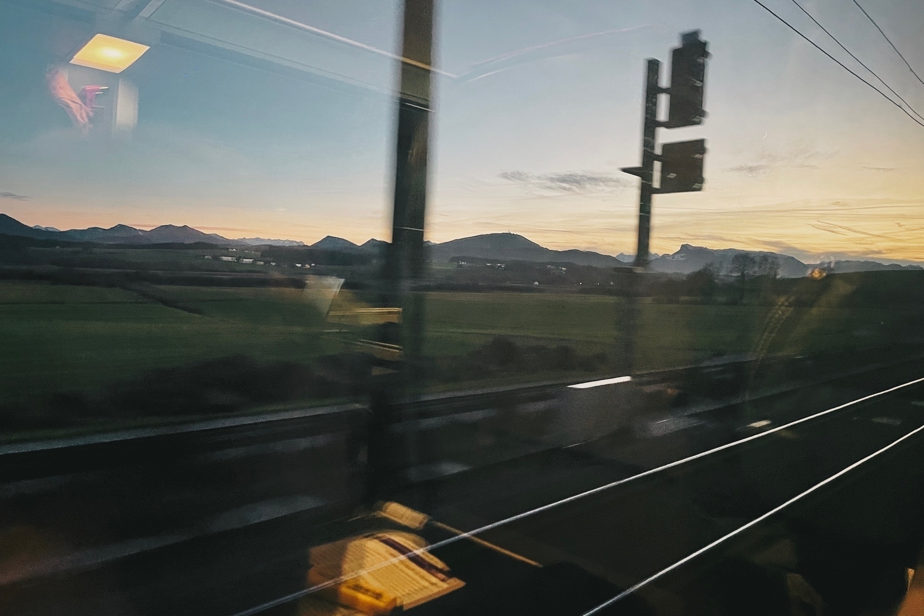 Taken from a moving train you see mountains in the distance, a bit after sundown; in the foreground you see train tracks and signs and in the reflection of the glass a book and pen