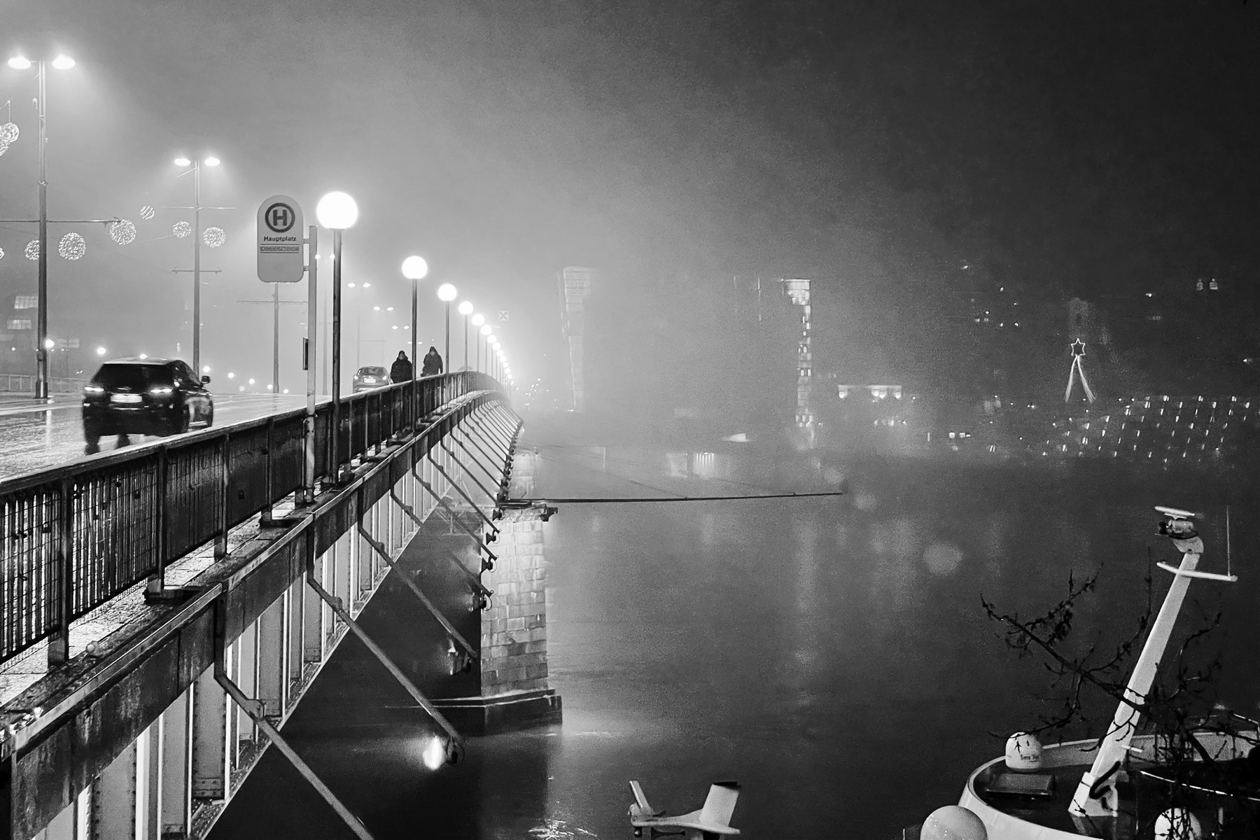 Black and white photo, a bridge with a car on it in the left side, the right shows the Danube at night and an illuminated building in the background; it is foggy