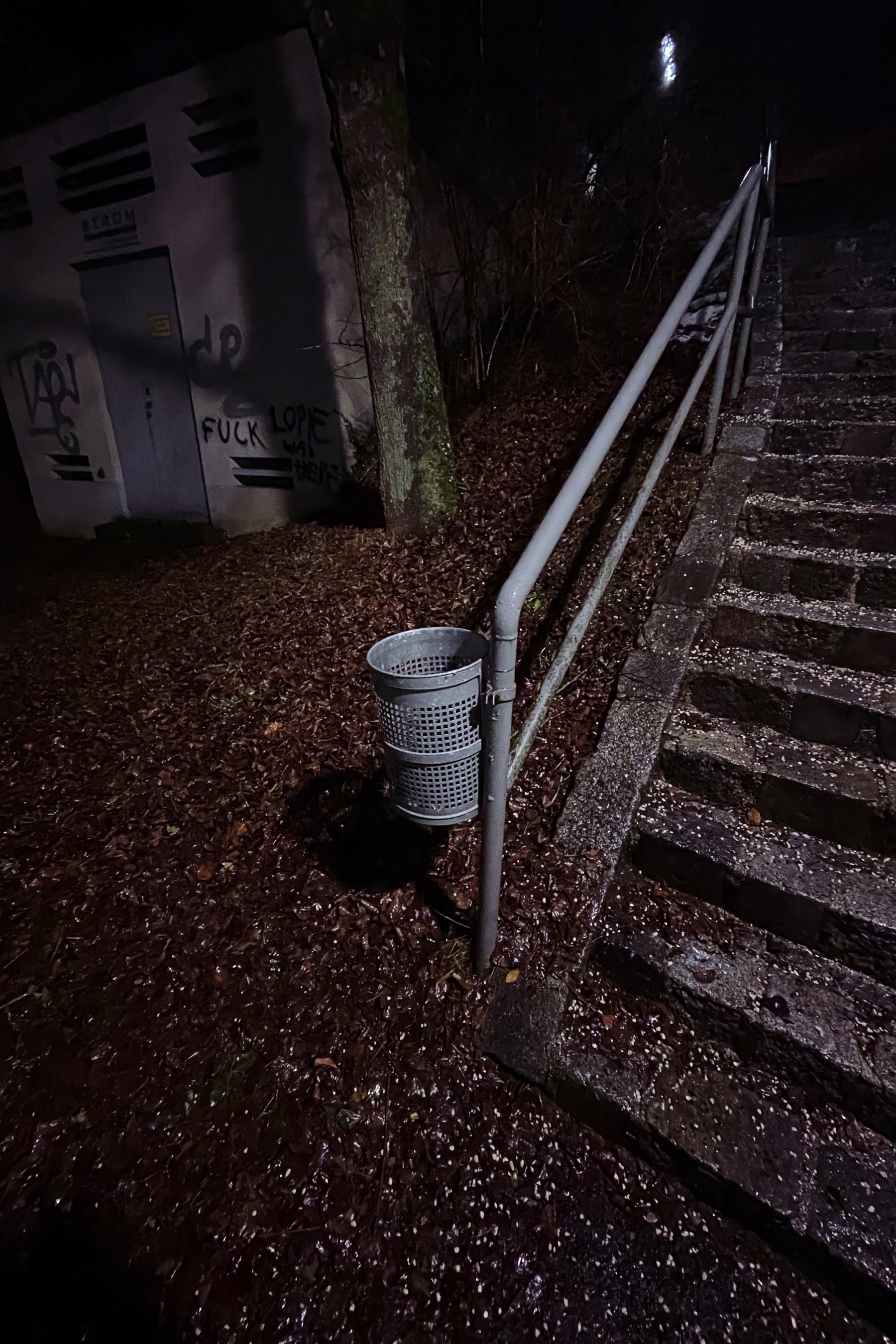 trash can fixed to the railing if a stair, illuminated by a street light. in the background there is a concrete building with graffiti 